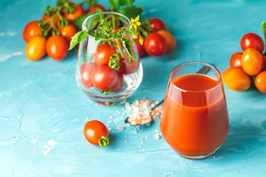 Glasses of fresh delicious jummy red tomato juice and fresh raw tomatoes with pink salt in spoon on light concrete surface. Close up. Gmo free. Natural good food.