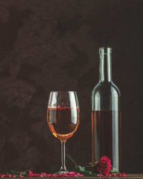 Bottle of rose wine and glass served with rose wine and rose petals, pink rose on dark background. Beautiul valentine or wedding greeting card.