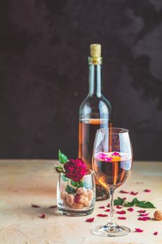 Bottle of rose wine and glass served with rose wine and rose petals, rose and candied fruit in glass on pink concrete table and dark background. Beautiful valentine or wedding greeting card.