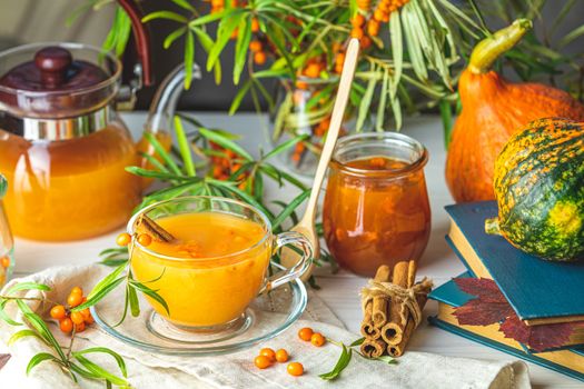 Cup and teapot of hot spicy tea with sea buckthorn, jam in the glass jar, branches of fresh berries on light woden table surface in the rustic room.