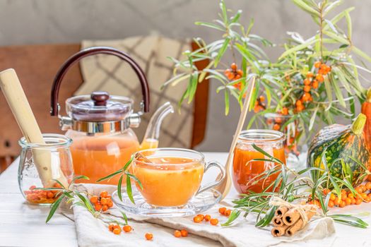 Cup and teapot of hot spicy tea with sea buckthorn, jam in the glass jar, branches of fresh berries on light woden table surface in the rustic room.