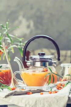 Cup and teapot of hot spicy tea with sea buckthorn, jam in the glass jar, branches of fresh berries on light woden table surface in the rustic room.