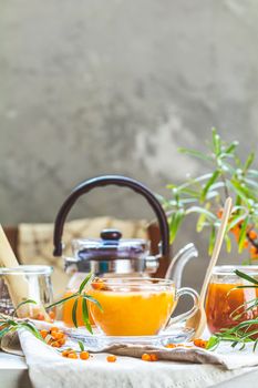 Cup and teapot of hot spicy tea with sea buckthorn, jam in the glass jar, branches of fresh berries on light woden table surface in the rustic room