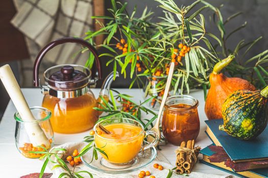 Cup and teapot of hot spicy tea with sea buckthorn, jam in the glass jar, branches of fresh berries on light woden table surface in the rustic room