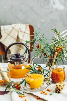 Cup and teapot of hot spicy tea with sea buckthorn, jam in the glass jar, branches of fresh berries on light woden table surface in the rustic room