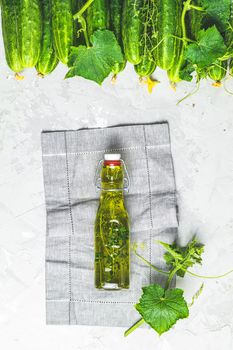 Bottle of olive oil with thyme on linen cloth with green fresh cucumbers with leaves and branches over gray texture background. Flat lay, space. Autumn home harvesting.