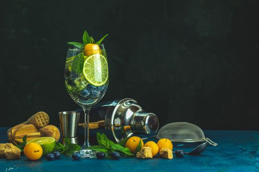 Summer alcoholic cocktail blueberry mojito with rum, mint, lime and ice, yellow cherry plums, bar tools, dark background, selective focus, copy space