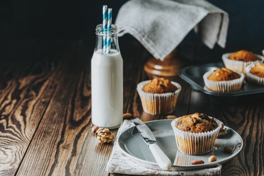 Vanilla caramel muffins in paper cups and bottles of milk on dark wooden background. Delicious cupcake with raisins, almonds and nuts. Homemade biscuit cakes. Copy space for text.