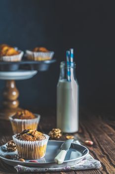 Delicious cupcake with raisins, almonds and nuts. Homemade biscuit cakes. Vanilla caramel muffins in paper cups and bottles of milk on dark wooden background. Copy space for text.