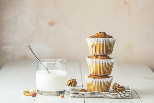 Vanilla caramel muffins in paper cups and glass of milk on white wooden background. Delicious cupcake with raisins, almonds and nuts. Homemade biscuit cakes. Copy space for text.