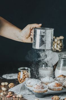 Sifting powdered sugar on delicious cupcake with raisins, almonds and nuts in kitchen. Aged female hand sifting powdered sugar by sieve over vanilla caramel muffins in paper cups in bakeware. 