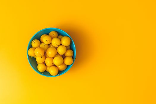 Creative summer pattern made of fresh yellow cherry plums in blue bowl on pastel yellow background. Fruit minimal concept. Flat lay.