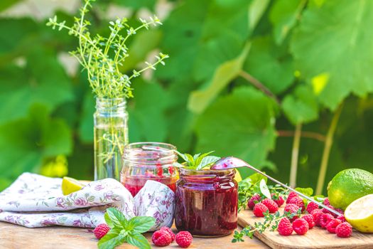Raspberry jam and fresh raspberry on a rustic wooden table outdoors. Sunny day, green leaves background