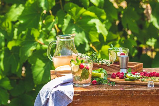 Refreshing mint cocktail mojito with rum, lime and raspberry, cold drink or beverage on a rustic wooden table outdoors. Sunny day, green leaves background