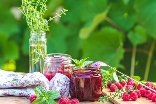 Raspberry jam and fresh raspberry on a rustic wooden table outdoors. Sunny day, green leaves background