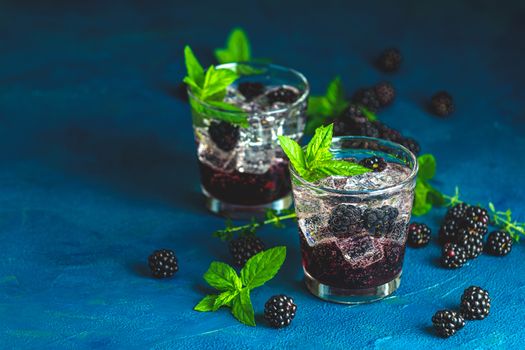 Cold summer berry drink with blackberries. Refreshing summer drink with syrup, blackberry and ice on dark blue concrete background