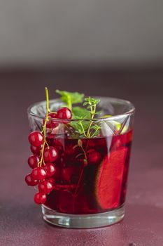 Glass of cold red cocktail with currant, lime, mint, thyme and ice in glass on dark red concrete surface table. Summer drinks and alcoholic cocktails. Alcoholic cocktail Currant mojito.