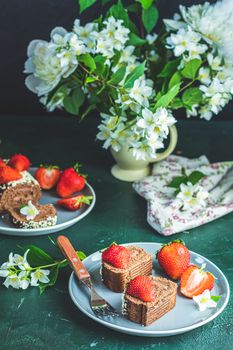 Chocolate rollcake with fresh strawberries in ceramic plate, jasmine and white peonies on dark green concrete surface table