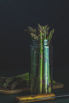 Green asparagus in glass jar standing on table at dark wall background. Fresh asparagus store. Spring seasonal vegetables. Copy space 