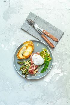 Green asparagus and boiled poached egg with hamon (jamon) and chopped parmigiano (parmesan) on a light gray plate over concrete background