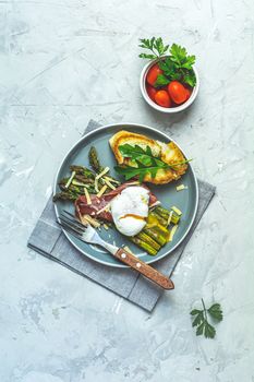 Green asparagus and boiled poached egg with hamon (jamon) and chopped parmigiano (parmesan) on a light gray plate over concrete background