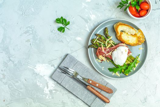 Green asparagus and boiled poached egg with hamon (jamon) and chopped parmigiano (parmesan) on a light gray plate over concrete background