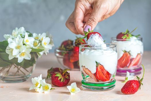 Woman hand holding cream strawberry. Glass bowl of strawberries with whipped cream and mint. Jasmine flowers. Concrete surface, copy space  for you text.
