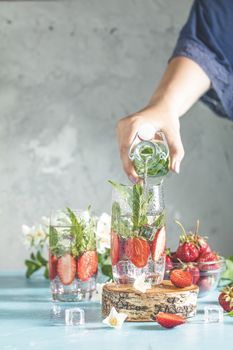 Women hand is pouring detox water from glass bottle to glass with strawberries, ice and mint. Summer fresh Mint soda cocktail, selective focus.