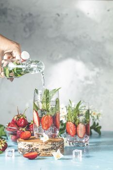 Women hand is pouring detox water from glass bottle to glass with strawberries, ice and mint. Summer fresh Mint soda cocktail, selective focus.