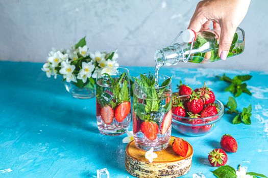 Woman pouring lemonade from bottle into glass. Detox infused water with strawberry and mint in highball glasses on blue concrete table background, copy space. Cold summer drink. Mineral water.