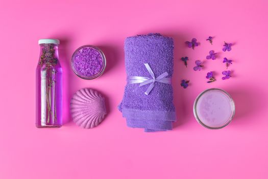 Spa products. Lavender bath salts, viola flowers, soap, cosmetic cream and towel. Violet purple concept. Flat lay on pink background, top view.