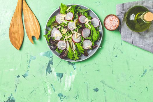 Salad with radishes, olive oil,  pink salt, empty ceramic bowl, wooden spoons for salad over green concrete concrete texture background. Top view, healthy food concept, top view, copy space for you text.