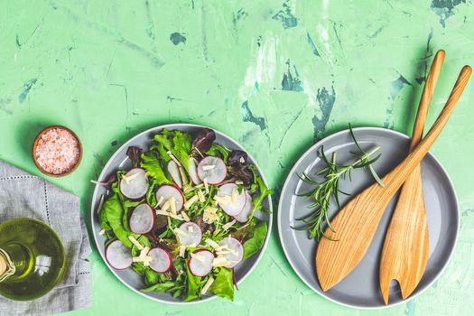 Salad with radishes, olive oil,  pink salt, empty ceramic bowl, wooden spoons for salad over green concrete concrete texture background. Top view, healthy food concept, top view, copy space for you text.