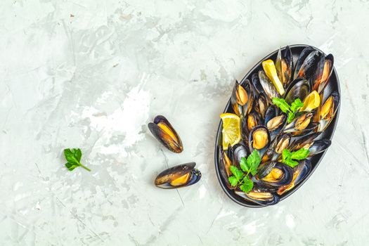 Seafood mussels with lemon and parsley in black metal plate on light gray concrete table surface, top view, copy space for you text