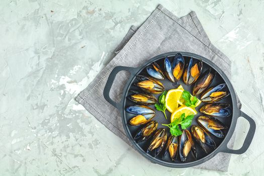 Seafood mussels with lemon and parsley in black metal pan on light gray concrete table surface, top view, copy space for you text