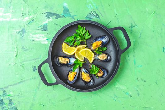 Seafood mussels with lemon and parsley in black metal pan on green concrete table surface, top view, copy space for you text