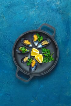 Seafood mussels with lemon and parsley in black metal pan on dark blue concrete table surface, top view, copy space for you text