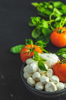 Mozzarella balls, buffalo in black ceramic plate, tomatoes and basil over dark background. With space. Rustic style. Ingredients for italian caprese salad