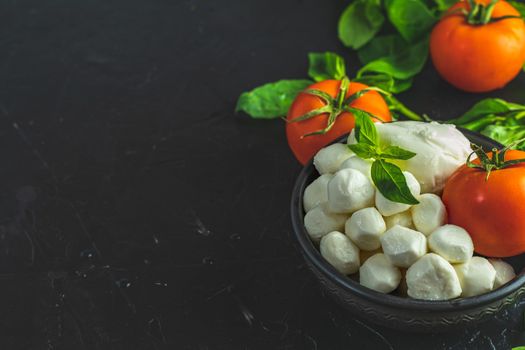Mozzarella balls, buffalo in black ceramic plate, tomatoes and basil over dark background. With space. Rustic style. Ingredients for italian caprese salad
