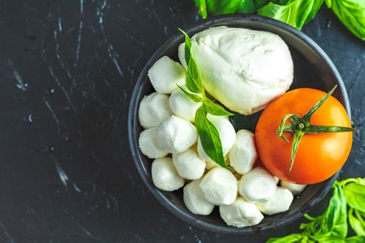 Mozzarella balls, buffalo in black ceramic plate, tomatoes and basil over dark background. With space. Rustic style. Ingredients for italian caprese salad. 