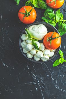 Mozzarella balls, buffalo in black ceramic plate, tomatoes and basil over dark background. With space. Rustic style. Ingredients for italian caprese salad. 