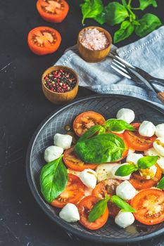 Italian caprese salad with sliced tomatoes, mozzarella cheese, basil and olive oil served in black ceramic plate with fork and knife on textile napkin over dark concrete table surface with copy space