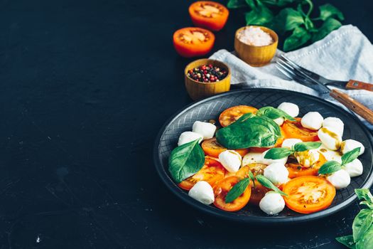 Italian caprese salad with sliced tomatoes, mozzarella cheese, basil and olive oil served in black ceramic plate with fork and knife on textile napkin over dark concrete table surface with copy space