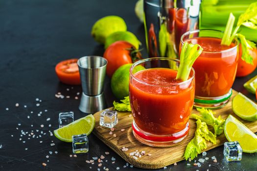 Tomato juice with celery, spices, salt and ice in portion glasses with copy space. Bloody Mary cocktail. Alcoholic drink and ingredients at dark concrete table surface. 