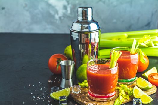 Tomato juice with celery, spices, salt and ice in portion glasses with copy space. Bloody Mary cocktail. Alcoholic drink and ingredients at dark concrete table surface. 