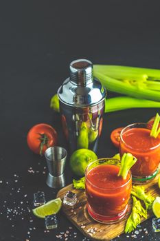 Tomato juice with celery, spices, salt and ice in portion glasses with copy space. Bloody Mary cocktail. Alcoholic drink and ingredients at dark concrete table surface. 