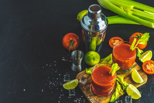Tomato juice with celery, spices, salt and ice in portion glasses with copy space. Bloody Mary cocktail. Alcoholic drink and ingredients at dark concrete table surface.