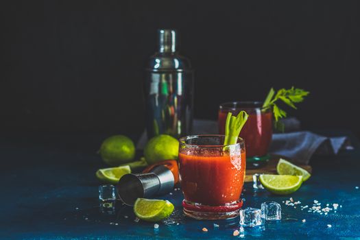 Tomato juice with celery, spices, salt and ice in portion drink glasses with copy space. Bloody Mary cocktail. Alcoholic drink and ingredients at dark blue concrete table surface.