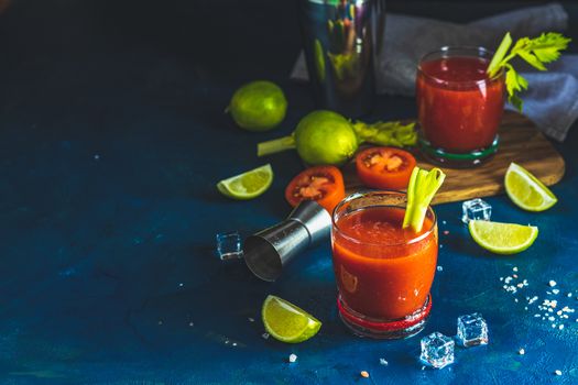Tomato juice with celery, spices, salt and ice in portion drink glasses with copy space. Bloody Mary cocktail. Alcoholic drink and ingredients at dark blue concrete table surface.