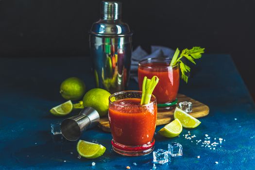 Tomato juice with celery, spices, salt and ice in portion drink glasses with copy space. Bloody Mary cocktail. Alcoholic drink and ingredients at dark blue concrete table surface.
